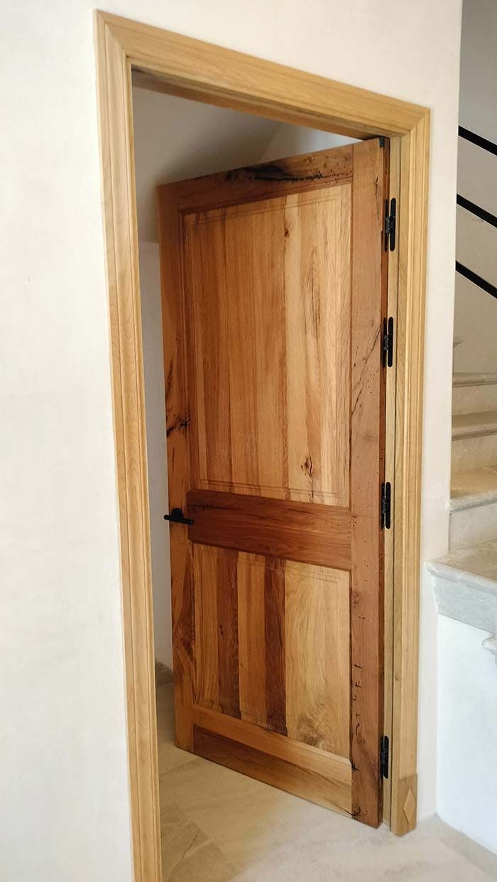 Puerta de madera, parcialmente abierta, dentro de un marco de madera de color claro, colocada en una pared de color claro.