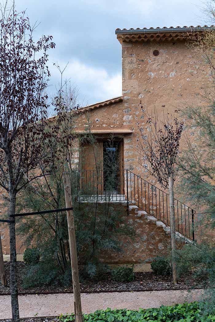 Edificio de piedra con escalera, puerta y pequeño balcón. Rodeado de árboles y arbustos, con un cielo nublado.