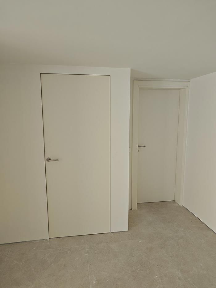 Dos puertas interiores blancas en una habitación con suelo gris claro. Una puerta es a ras de suelo y la otra está retranqueada.