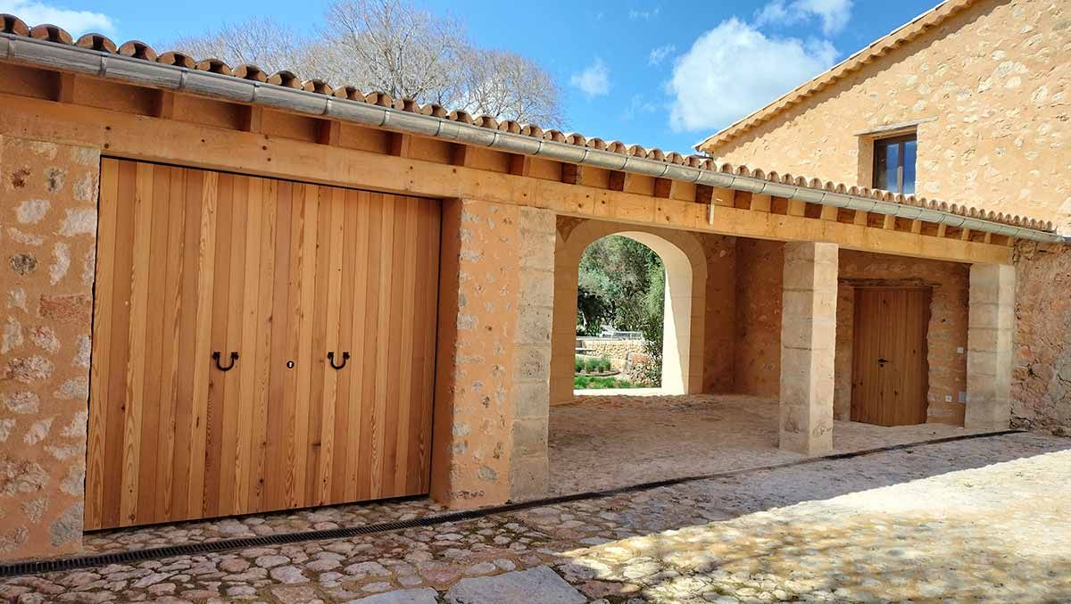 Puertas de garaje de madera y entrada arqueada bajo el alero de un edificio de piedra con techo de tejas.