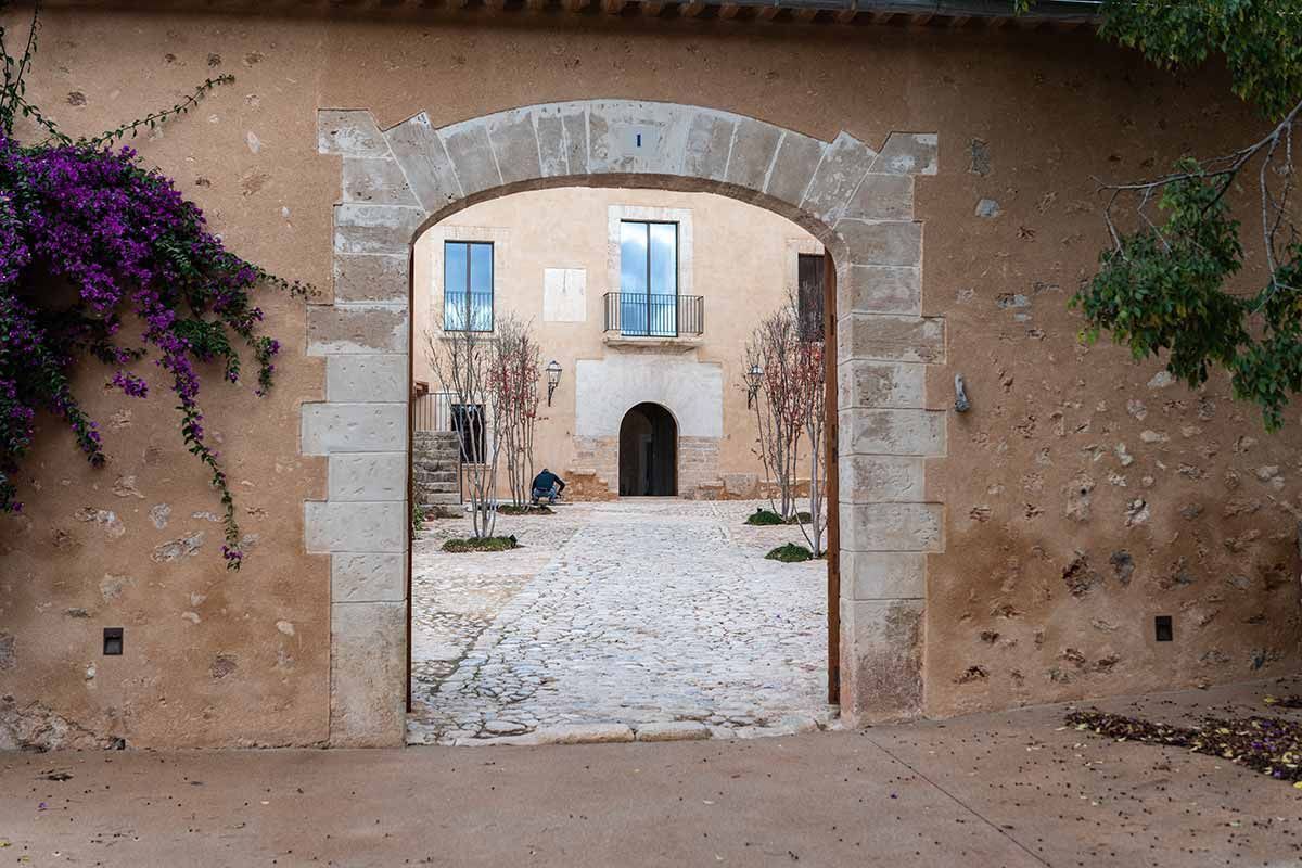 Arco de piedra que conduce a un patio adoquinado y a un edificio con balcón y ventanas.