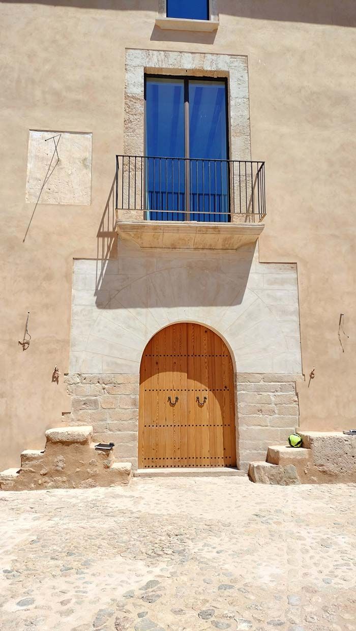Edificio beige con puerta de madera arqueada, balcón y escalones de piedra. El cielo azul se refleja en la ventana superior.