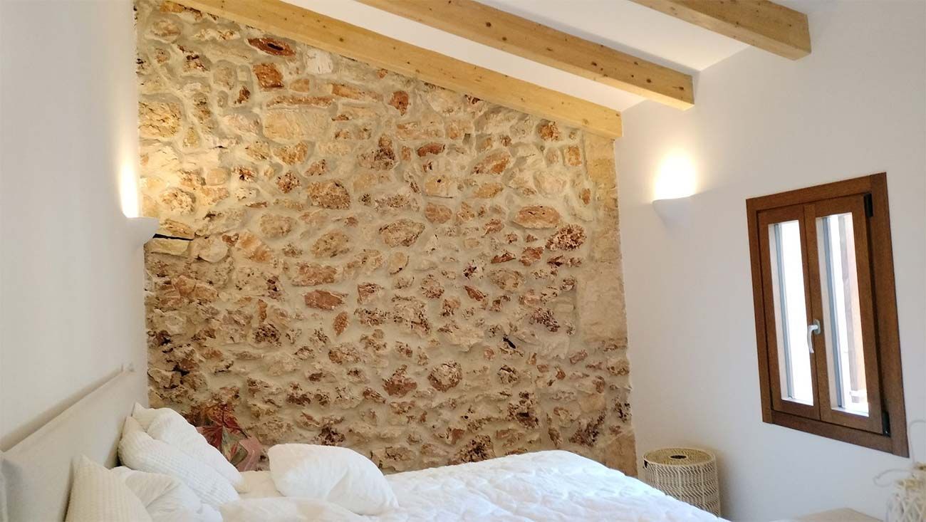 Dormitorio con pared con acento de piedra, ropa de cama blanca, vigas de madera, ventana pequeña y apliques de pared.
