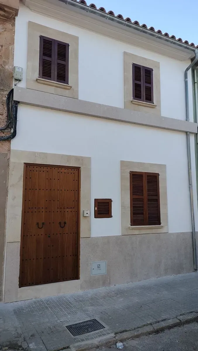 Edificio blanco de dos pisos con persianas y puerta marrones, molduras beige y una calle estrecha al frente.