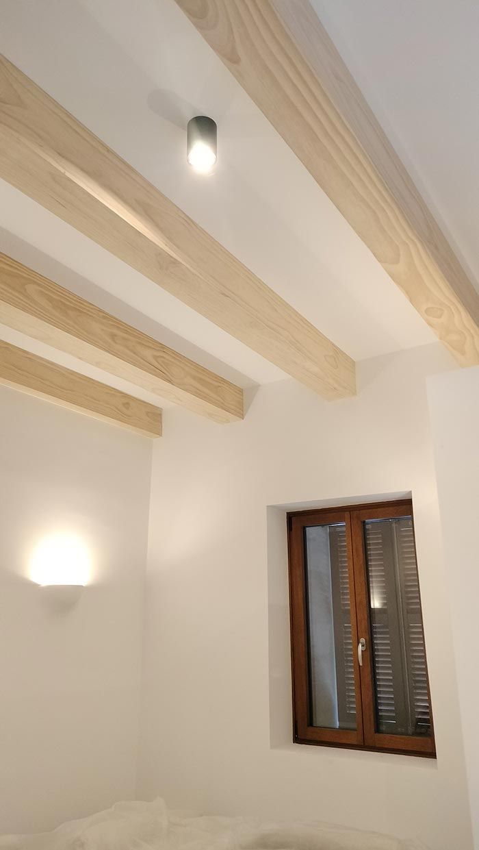 Habitación blanca con vigas y molduras de madera. Se ven una ventana y dos apliques.