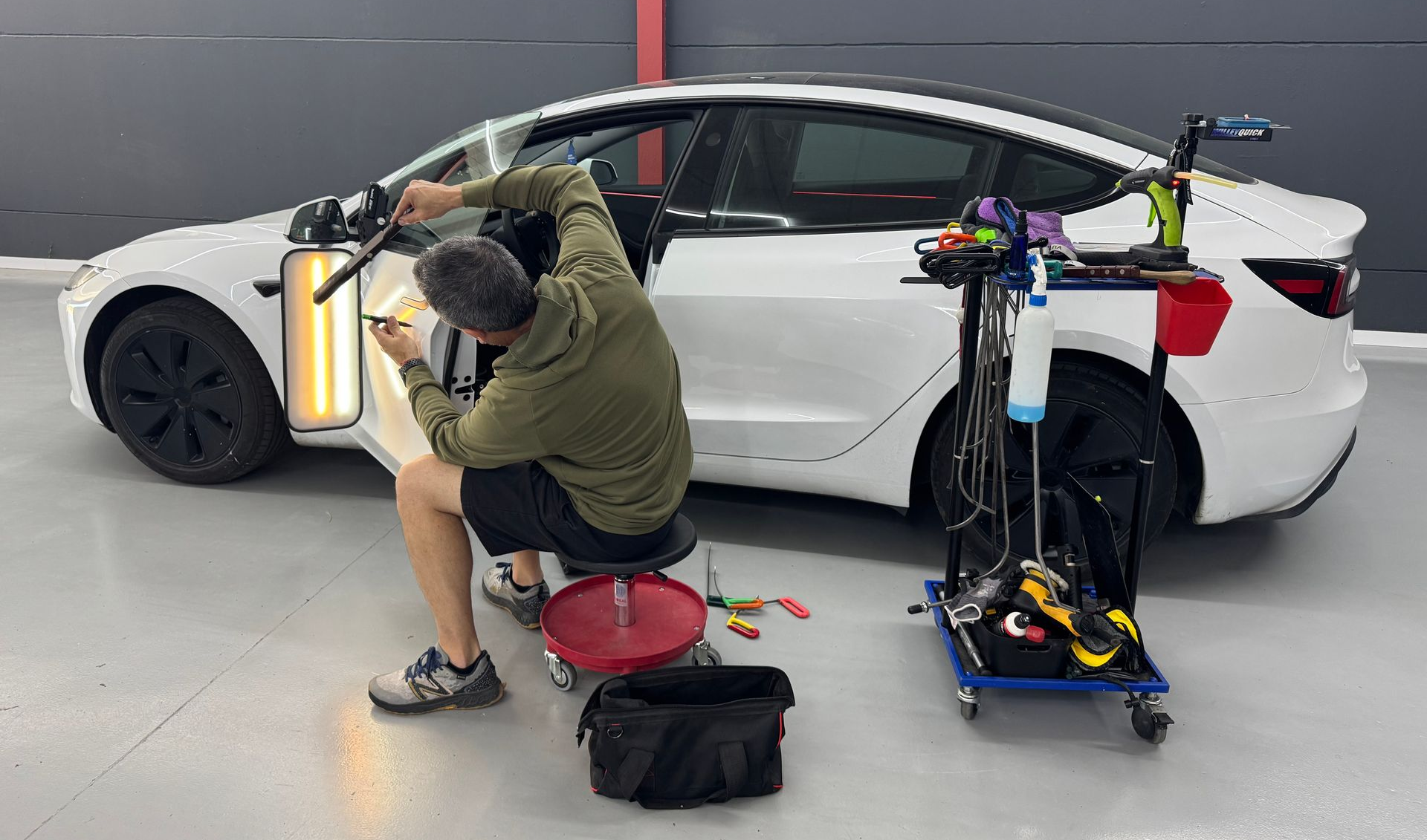 Persona trabajando en un sedán blanco en un garaje, rodeada de herramientas y equipos para la reparación de automóviles.