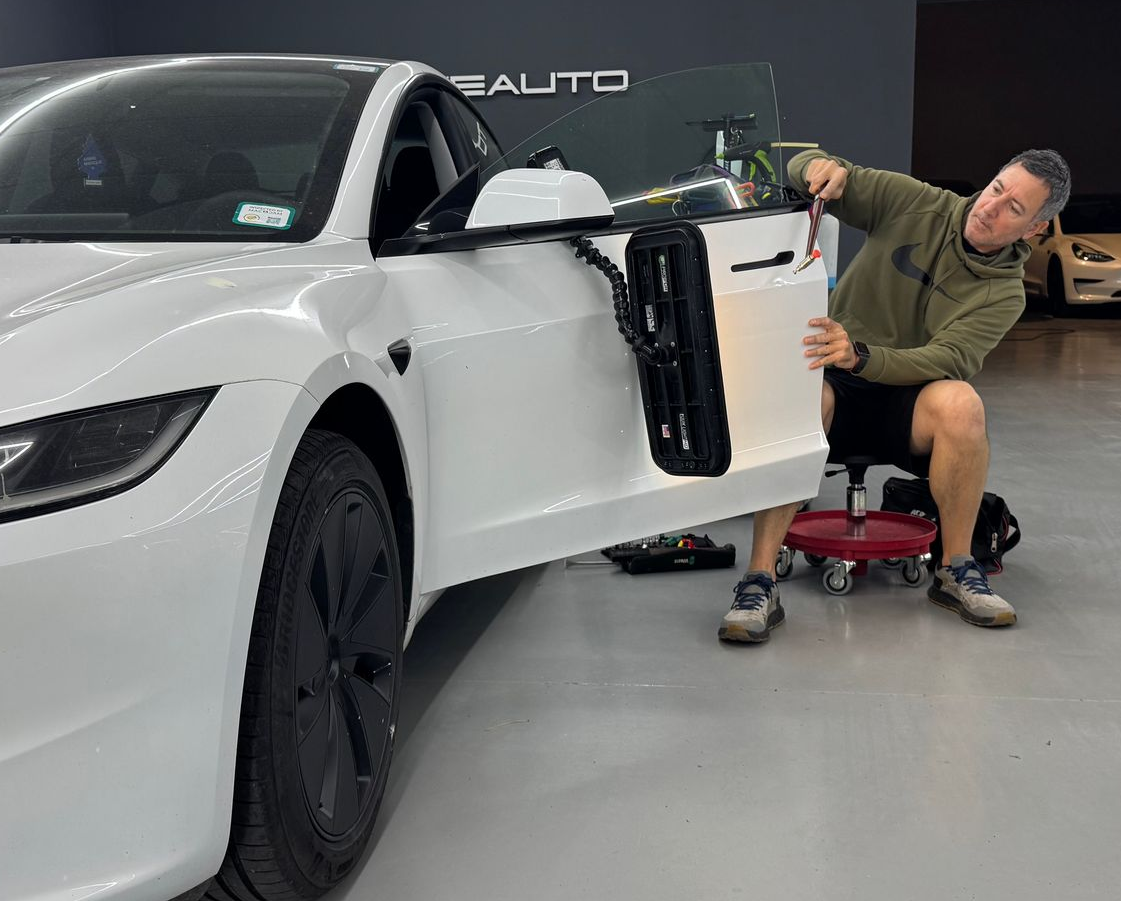 Un hombre sentado junto a un coche deportivo blanco, sujetando la puerta abierta en un garaje que parece una sala de exposición.