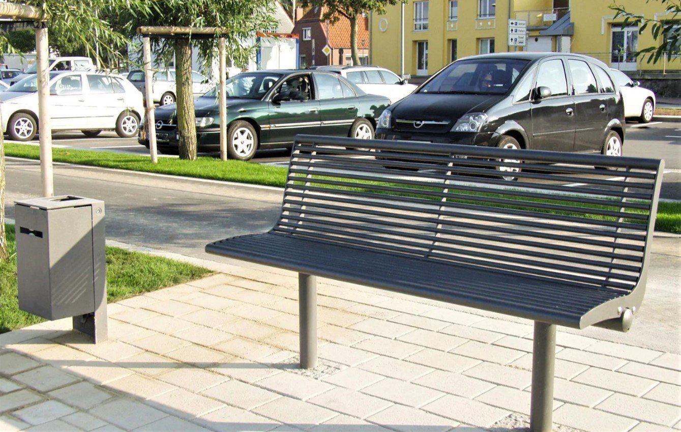 Tyhjä puistonpenkki roskakorin vieressä aurinkoisella jalkakäytävällä pysäköityjen autojen lähellä