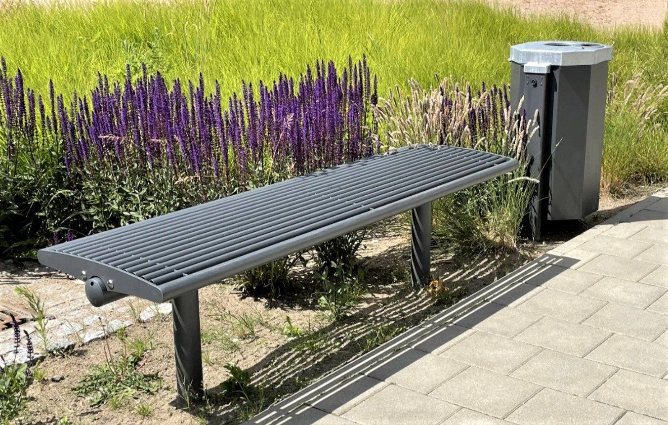 Metallinen penkki violettien kukkien ja roskakorin vieressä päällystetyllä polulla