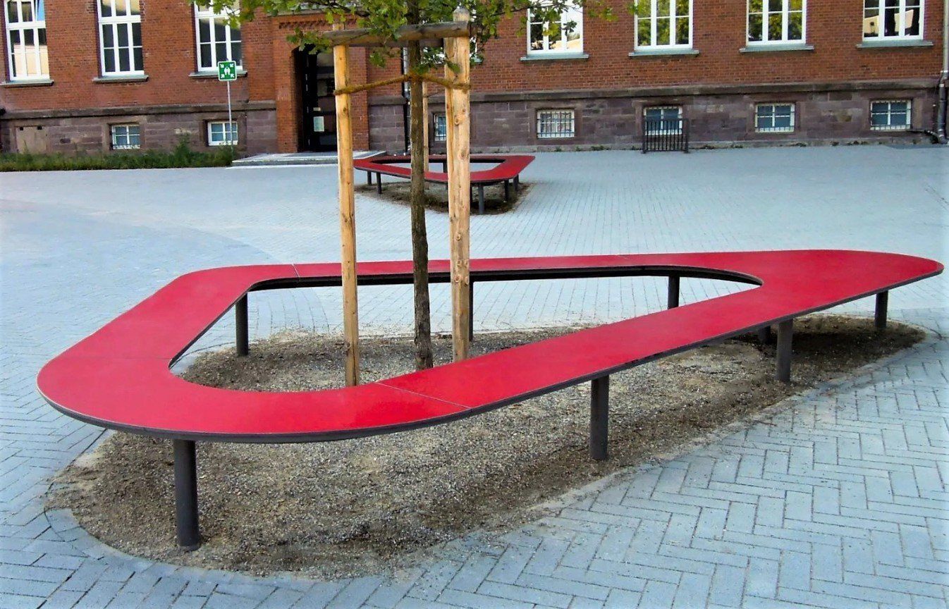 Punainen kaareva penkki puun ympärillä laatoitettulla sisäpihalla tiilirakennuksen ulkopuolella