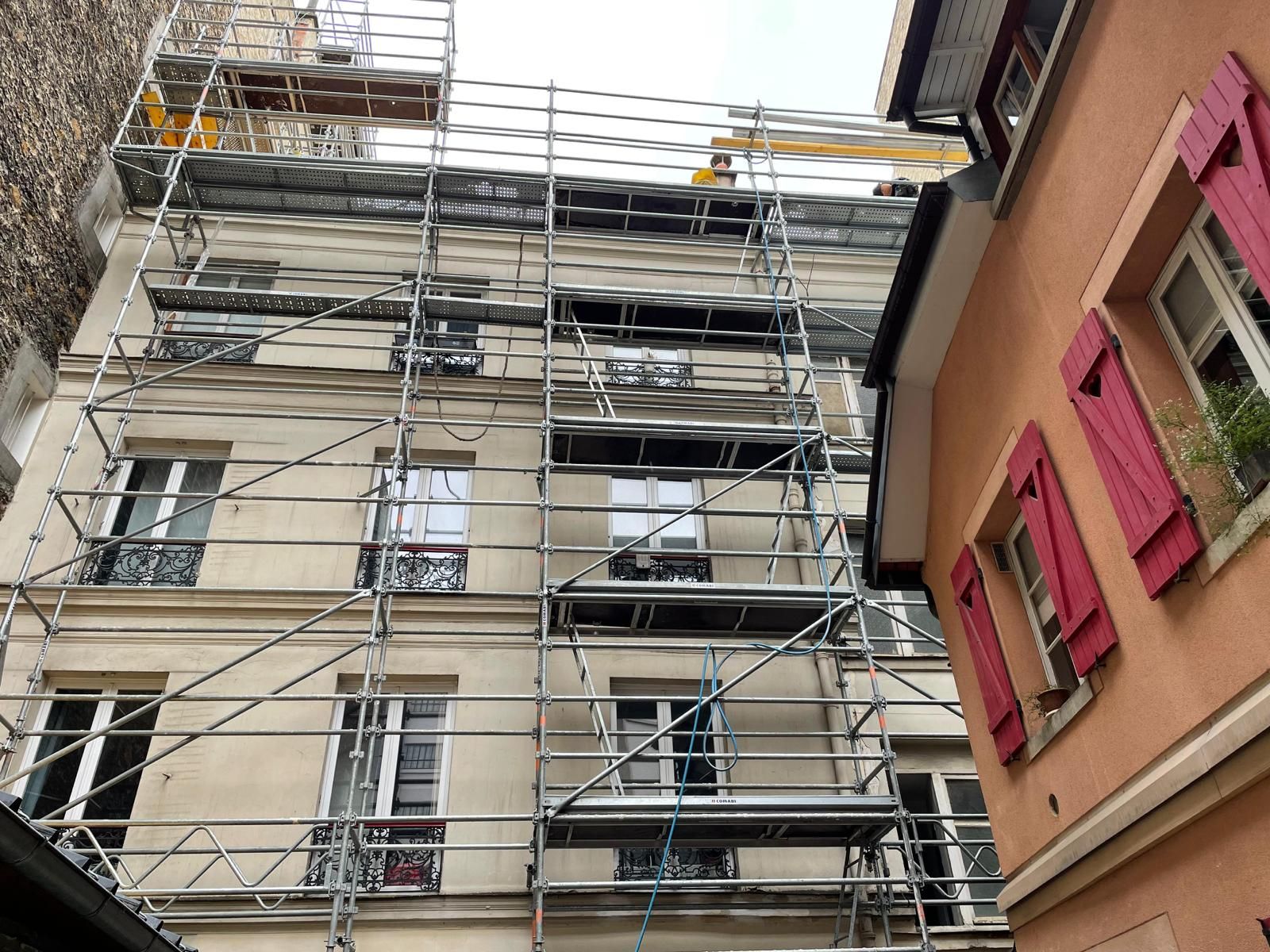 Façade d'un bâtiment en travaux, avec échafaudages. Volets rouges sur le bâtiment adjacent.