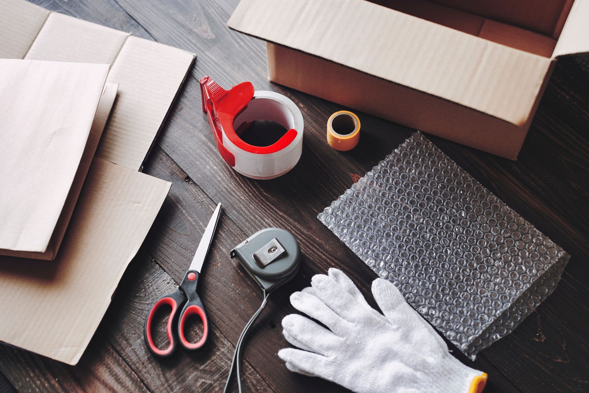 Une paire de ciseaux, des gants, un mètre, du papier bulle et du carton posés sur une table