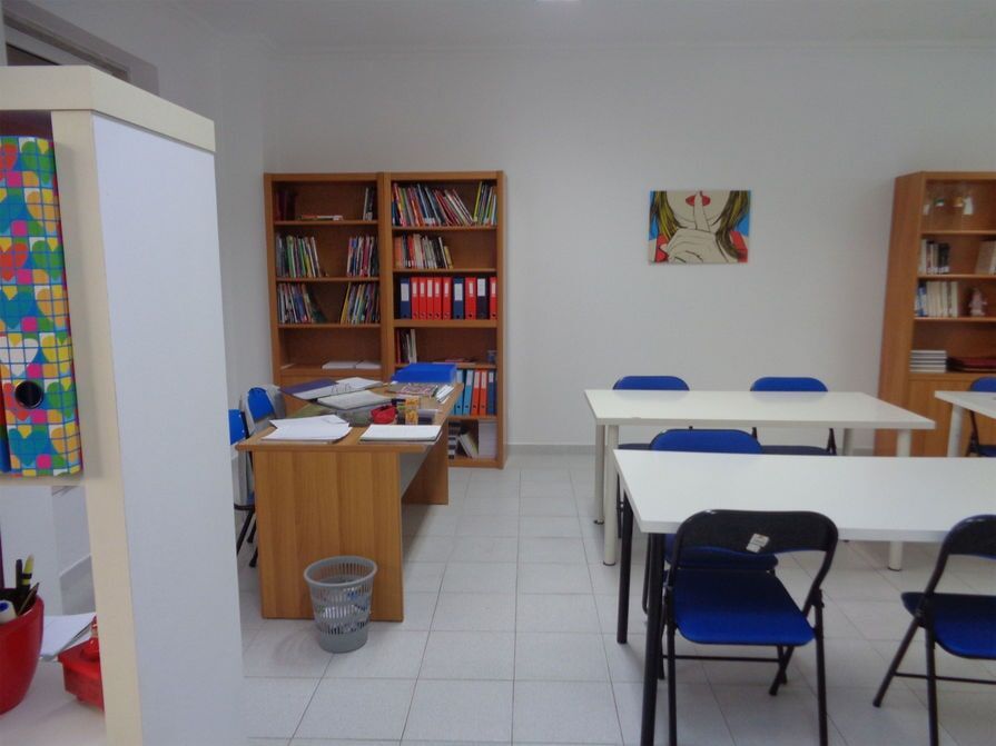 Sala de aula com carteiras, estantes, uma mesa para o professor e uma pintura colorida na parede.