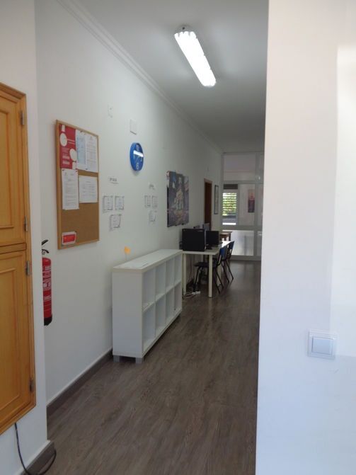 Corredor longo com paredes brancas, quadros de avisos e uma mesa com cadeiras no final.