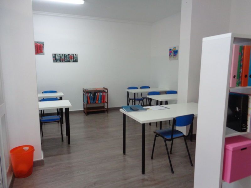 Sala de aula vazia com carteiras brancas e cadeiras azuis. Prateleiras com livros e pastas. Lixeira laranja.