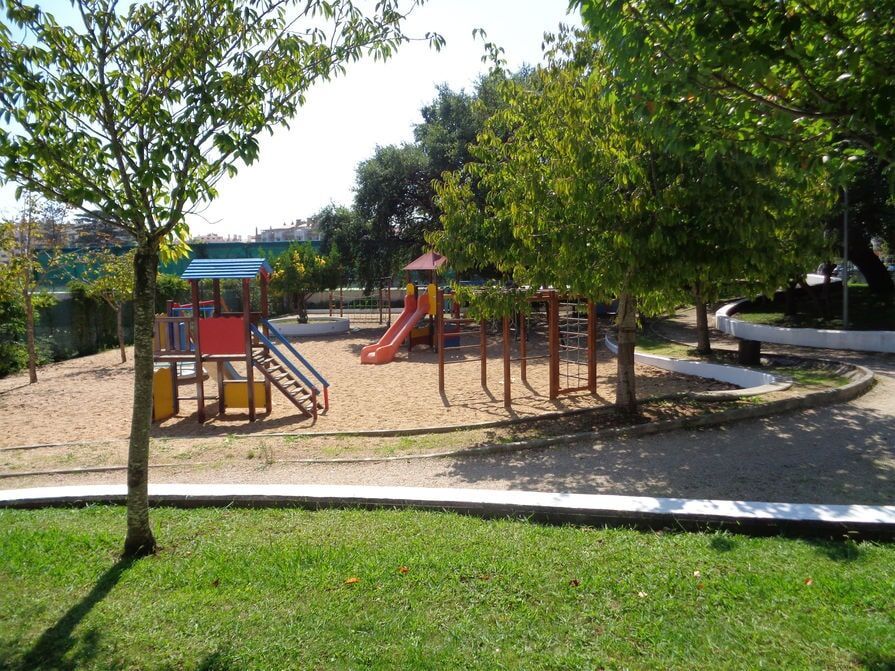 Parque infantil com estrutura de escalada, escorregador e balanços cercado por árvores e grama.