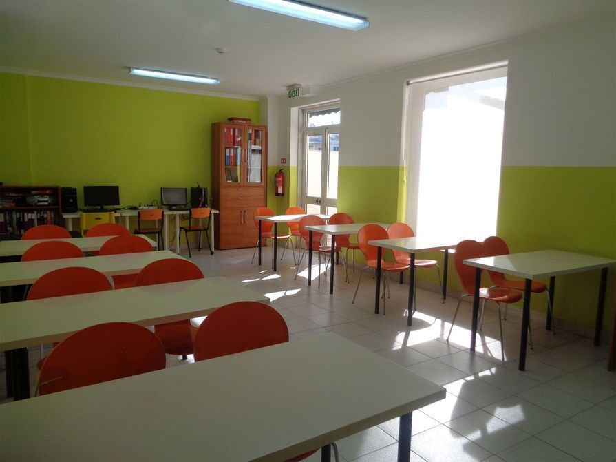 Sala de aula vazia com fileiras de mesas, cadeiras laranja, paredes verdes e brancas e uma janela iluminada.