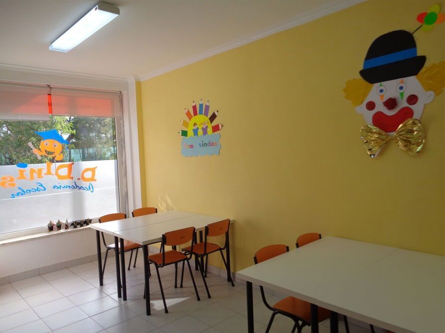 Uma sala de aula com paredes amarelas, mesas, cadeiras e decoração de palhaço. Luz do sol entrando pela janela.