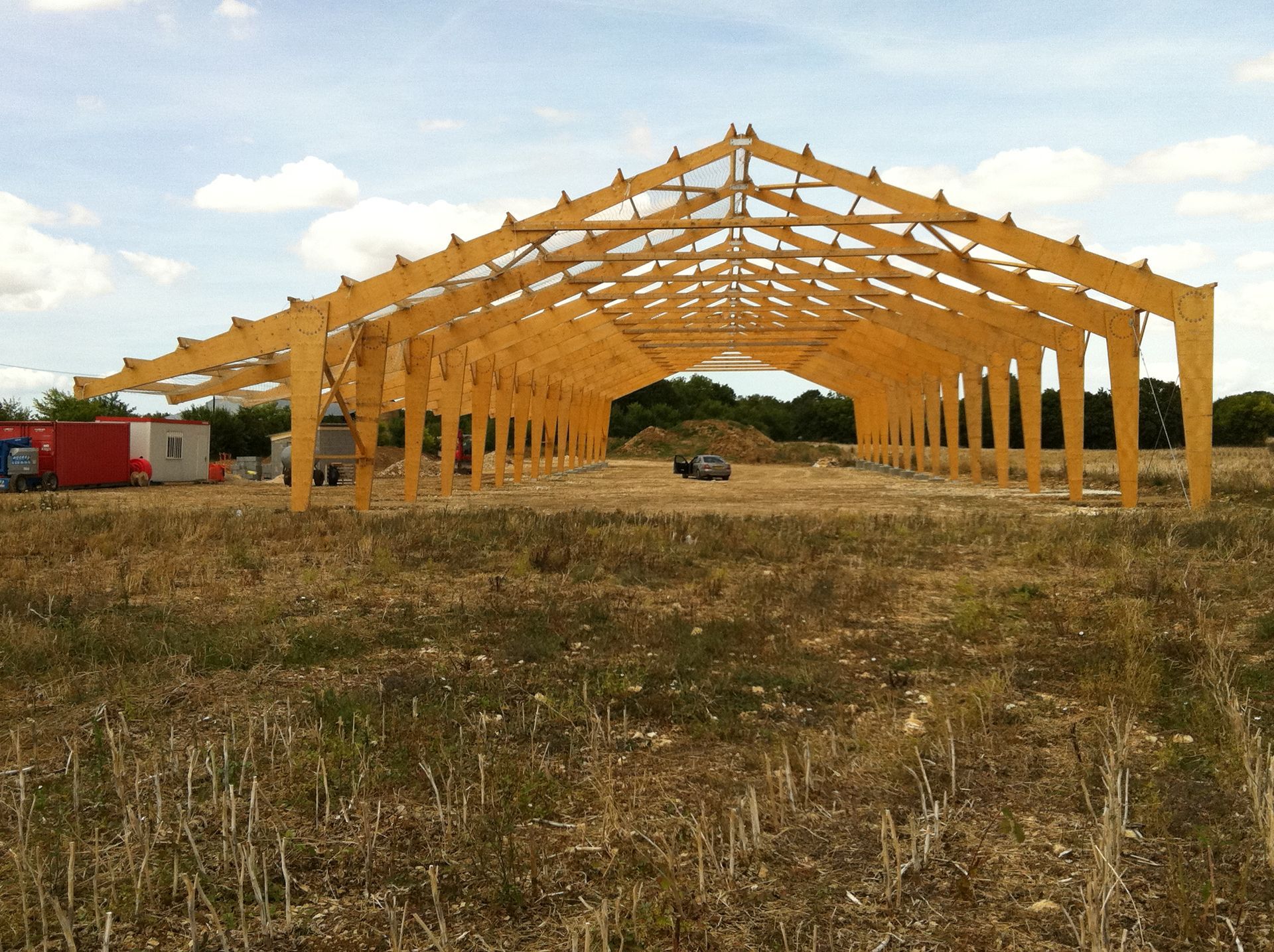 Charpente en bois d'un bâtiment dans un champ, sous un ciel nuageux.