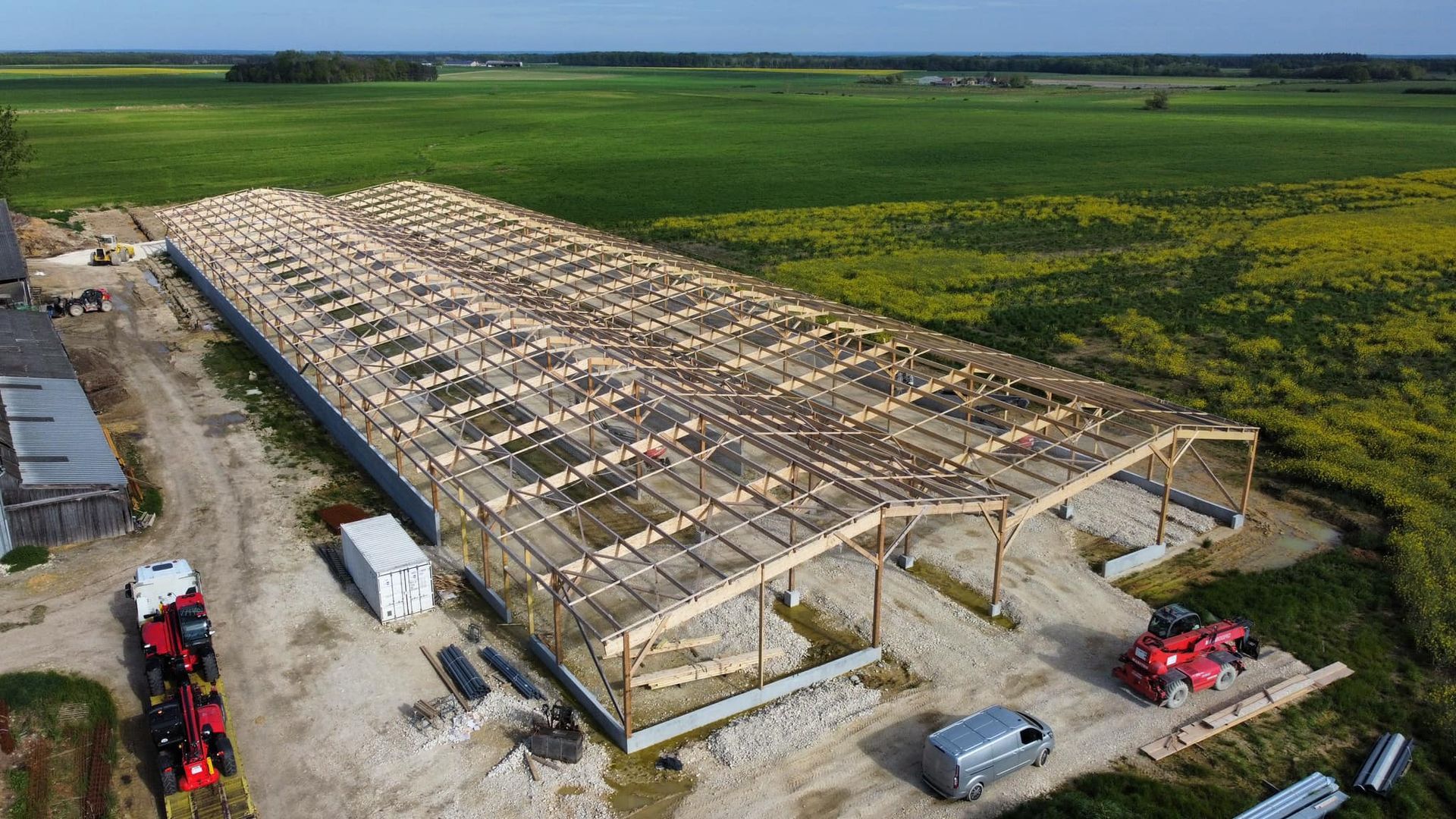 Vue aérienne d'une grange en construction. La toiture est soutenue par des poutres en bois. Des tracteurs et un champ se trouvent à proximité.