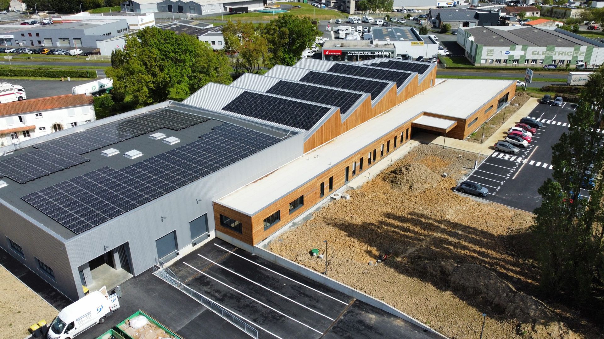 Vue aérienne d'un bâtiment moderne avec des panneaux solaires sur le toit, un parking et des arbres environnants.