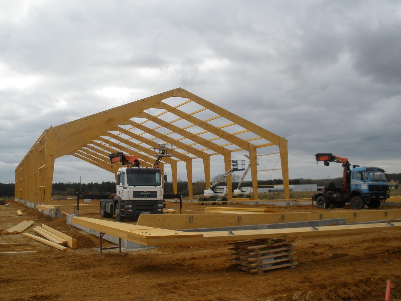 Construction d'un bâtiment à ossature bois avec camions et grue sur un chantier brun sous un ciel nuageux.