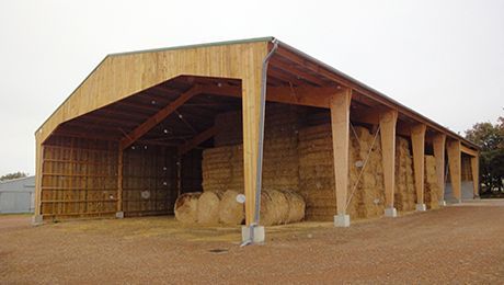 Grange à foin en bois remplie de bottes de foin, située à l'extérieur sur un terrain gravillonné.
