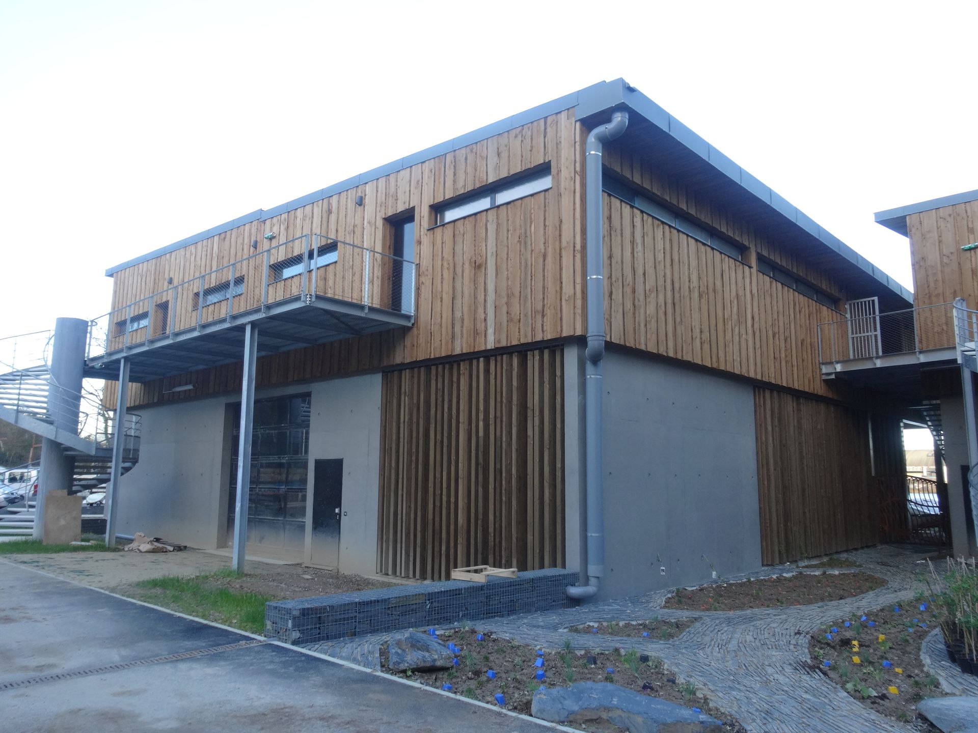 Bâtiment avec bardage en bois sur béton ; murs gris, balcon en métal, ciel nuageux.