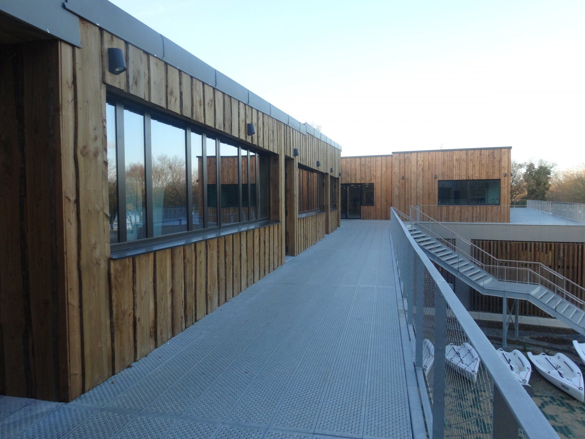 Façade en bois d'un bâtiment avec de hautes fenêtres, une allée et des barques en contrebas.