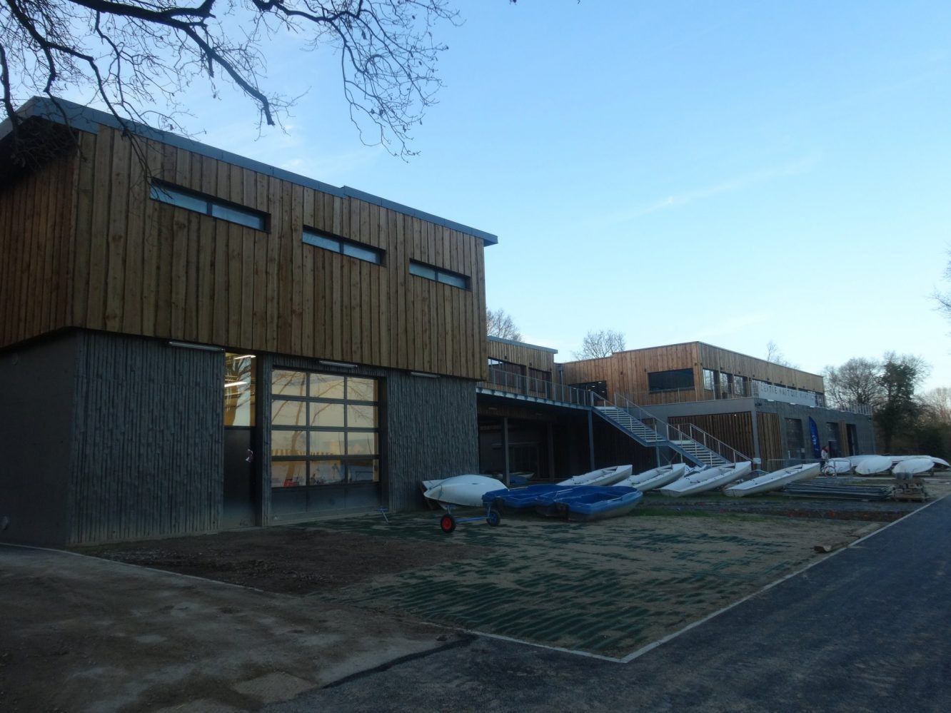 Hangar à bateaux moderne avec bardage en bois, donnant sur des bateaux sur l'herbe, par une journée ensoleillée.