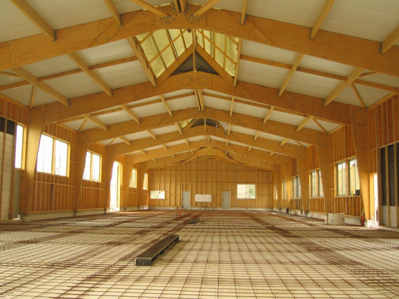 Intérieur d'un grand bâtiment en bois en construction, avec poutres apparentes, fenêtres et plancher en treillis métallique.