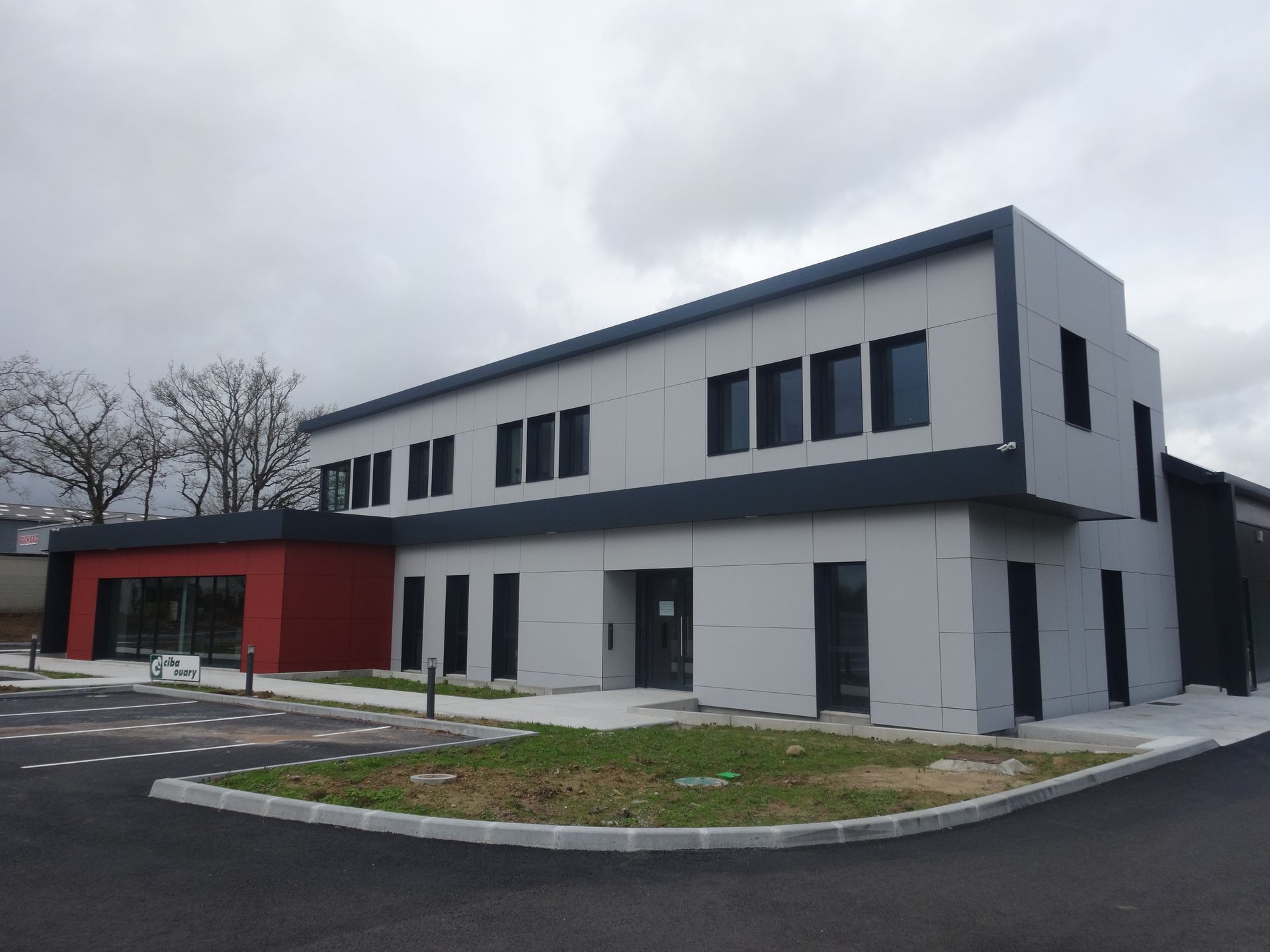 Bâtiment moderne gris et rouge aux finitions sombres, sous un ciel nuageux.