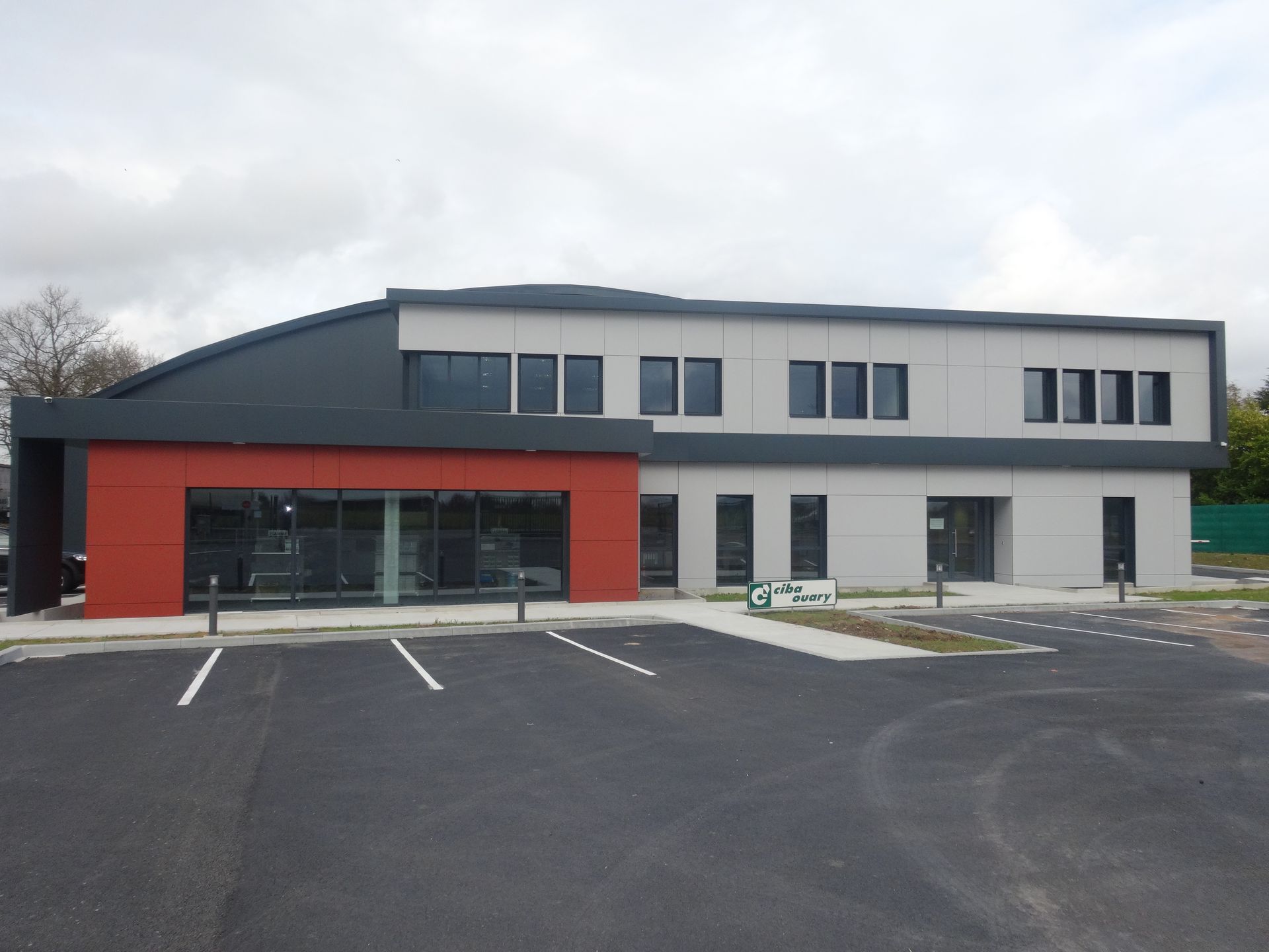 Bâtiment moderne de deux étages avec une façade rouge et grise, des fenêtres en verre et un parking pavé.