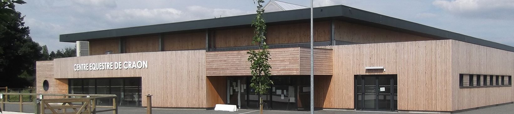 Un bâtiment en bois clair avec un toit sombre et un petit arbre devant. Ciel nuageux.