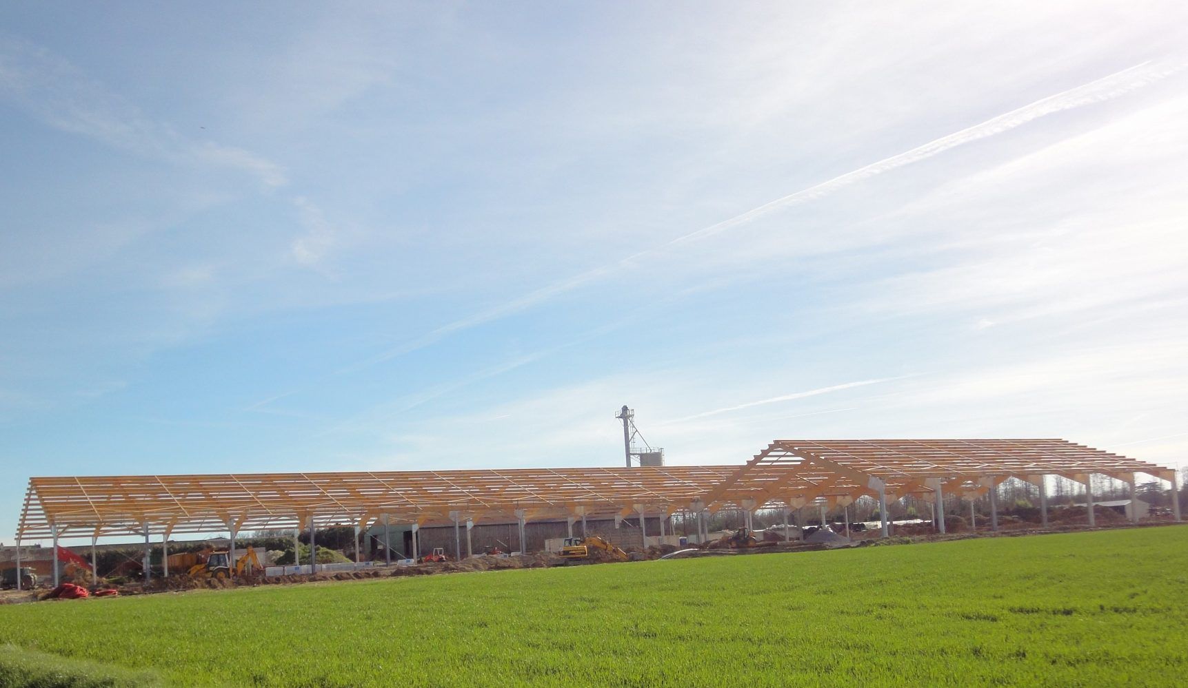 Construction d'un grand bâtiment aux poutres apparentes, situé dans un champ sous un ciel bleu.
