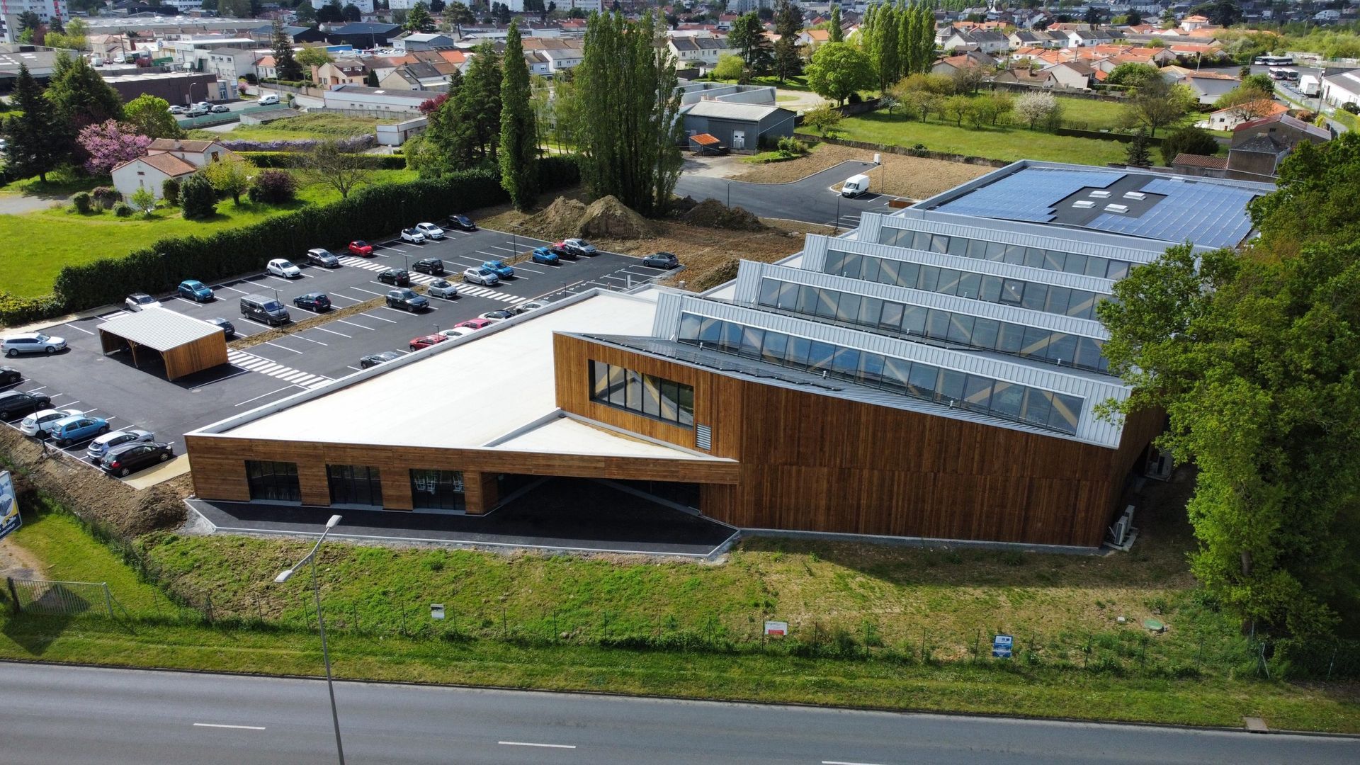 Bâtiment moderne avec toit en verre incliné et façade en bois, situé à proximité d'un parking et d'une route.