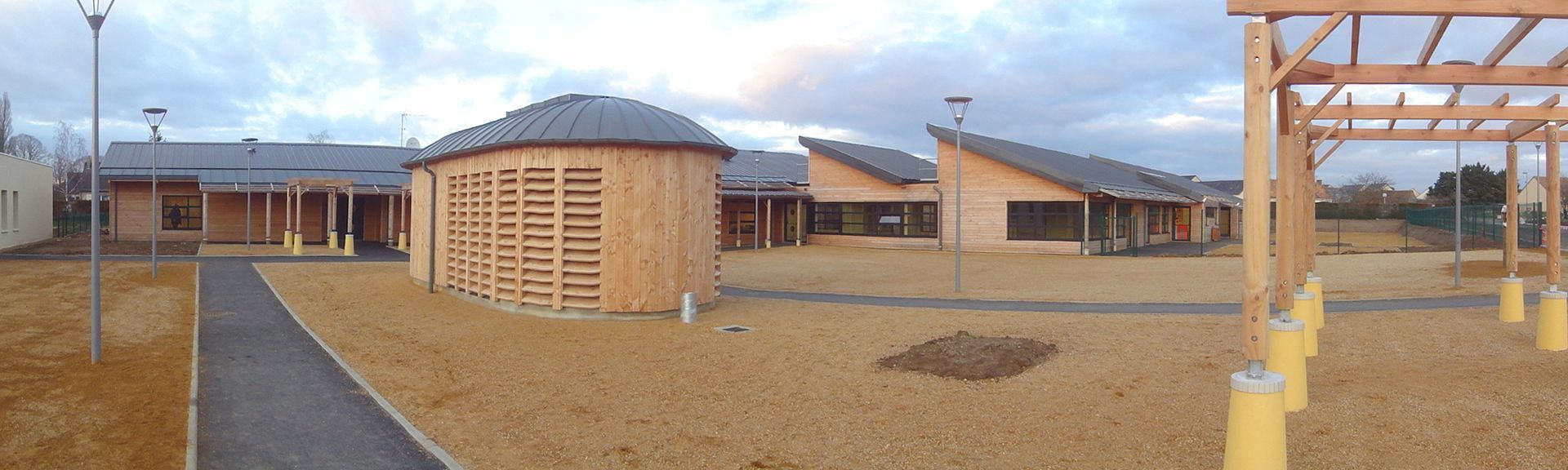 Vue extérieure d'une école avec un chemin, des structures en bois et un sol en gravier sous un ciel nuageux.