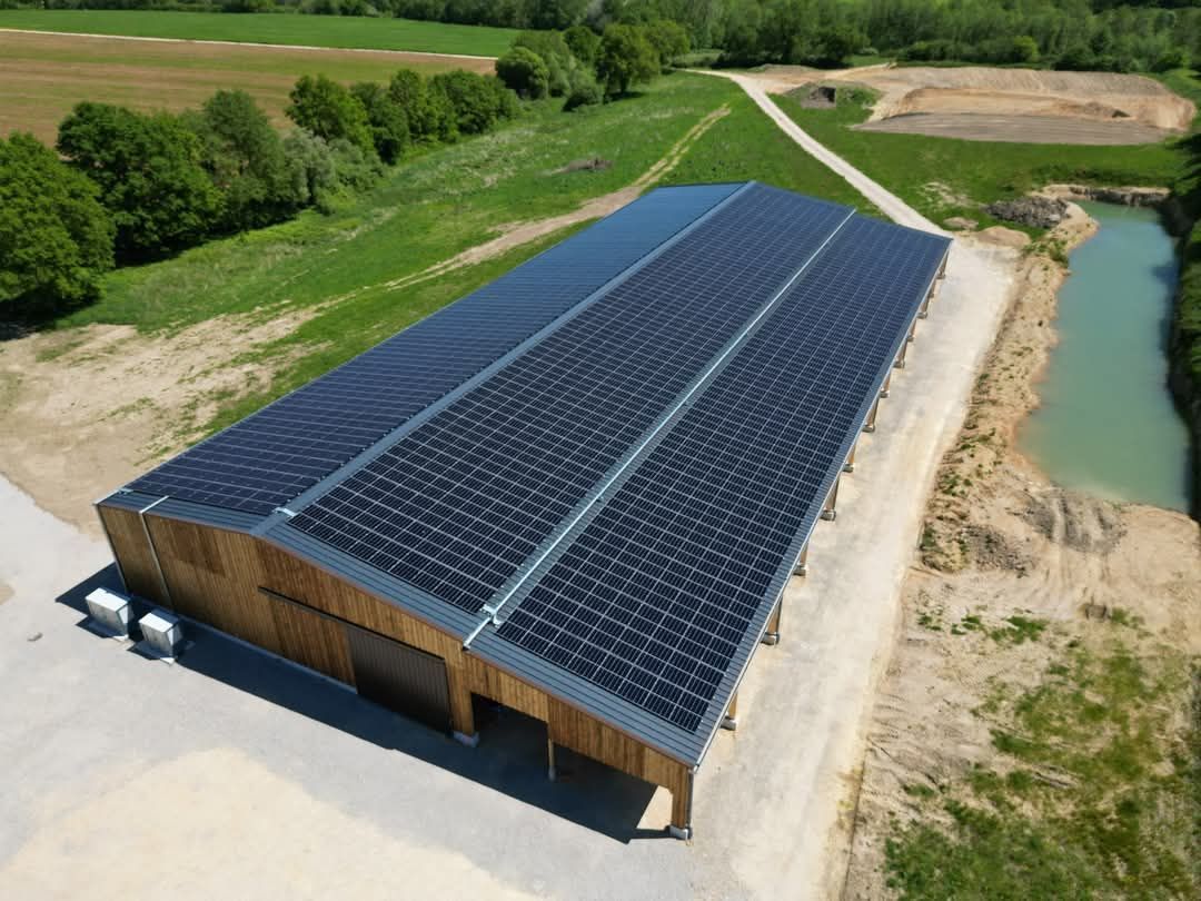 Grand bâtiment avec panneaux solaires sur le toit ; construction en bois et en pierre. Situé à proximité de champs verdoyants et d'un étang.