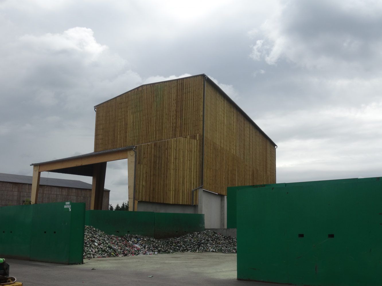 Bâtiment en bois avec abri attenant, barrières vertes, bouteilles et ciel nuageux.