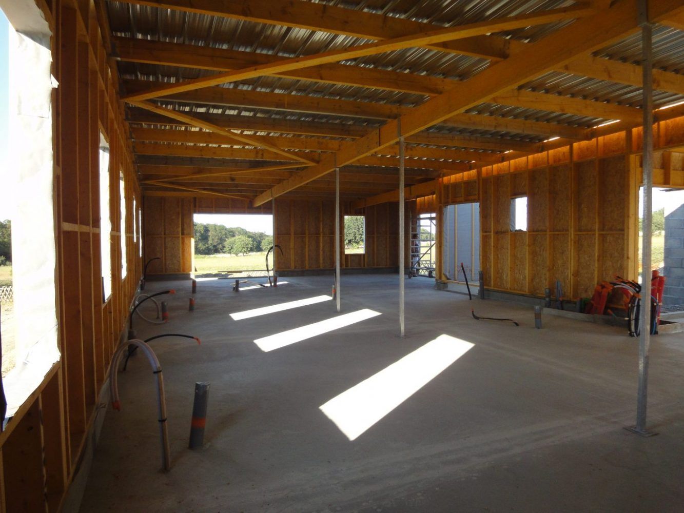 Intérieur d'un bâtiment en construction, montrant la charpente en bois, le sol en béton et la lumière du soleil qui entre par les fenêtres.
