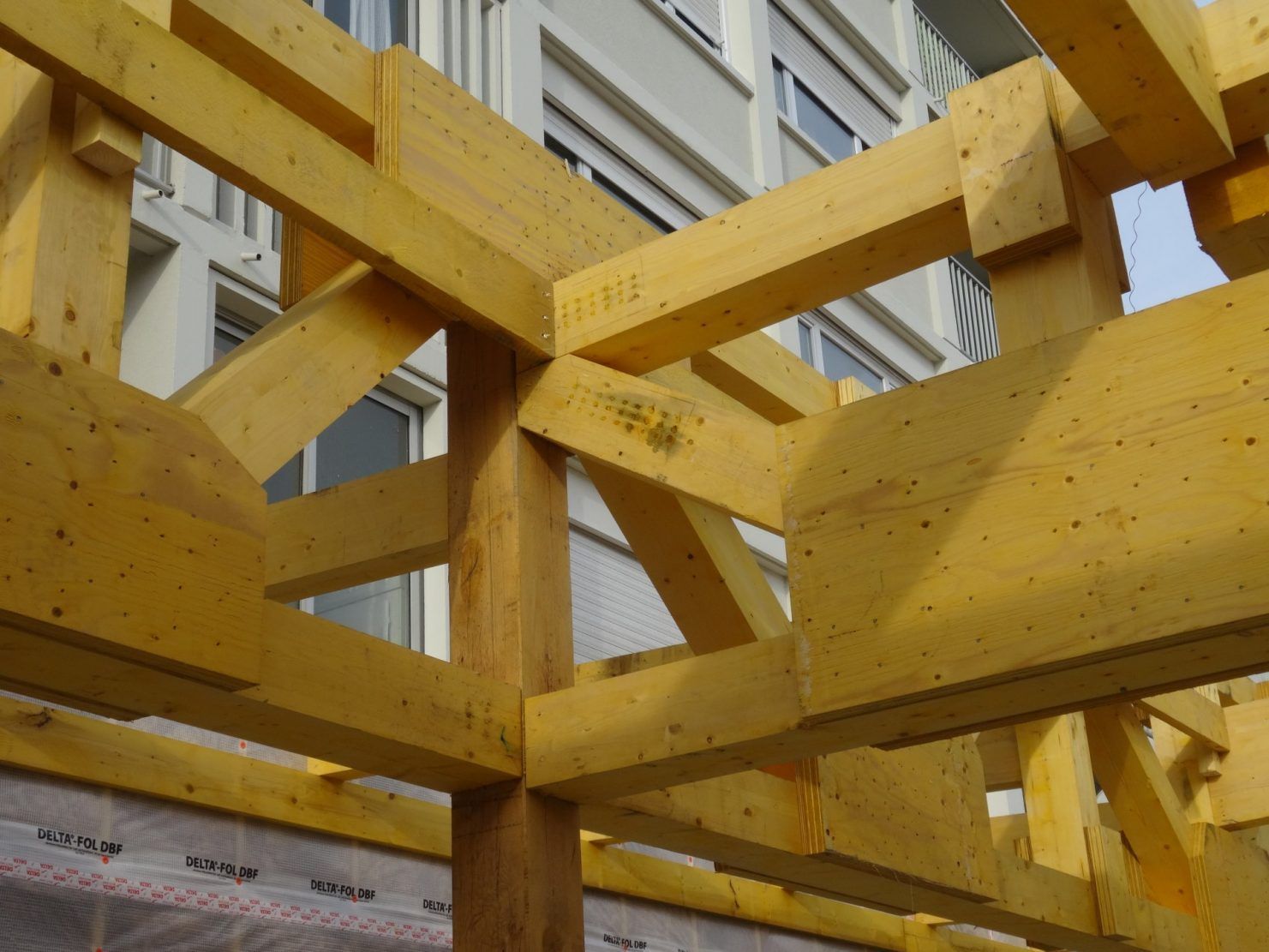 Des poutres en bois jaune forment une structure, probablement celle d'un bâtiment, avec un édifice urbain en arrière-plan.