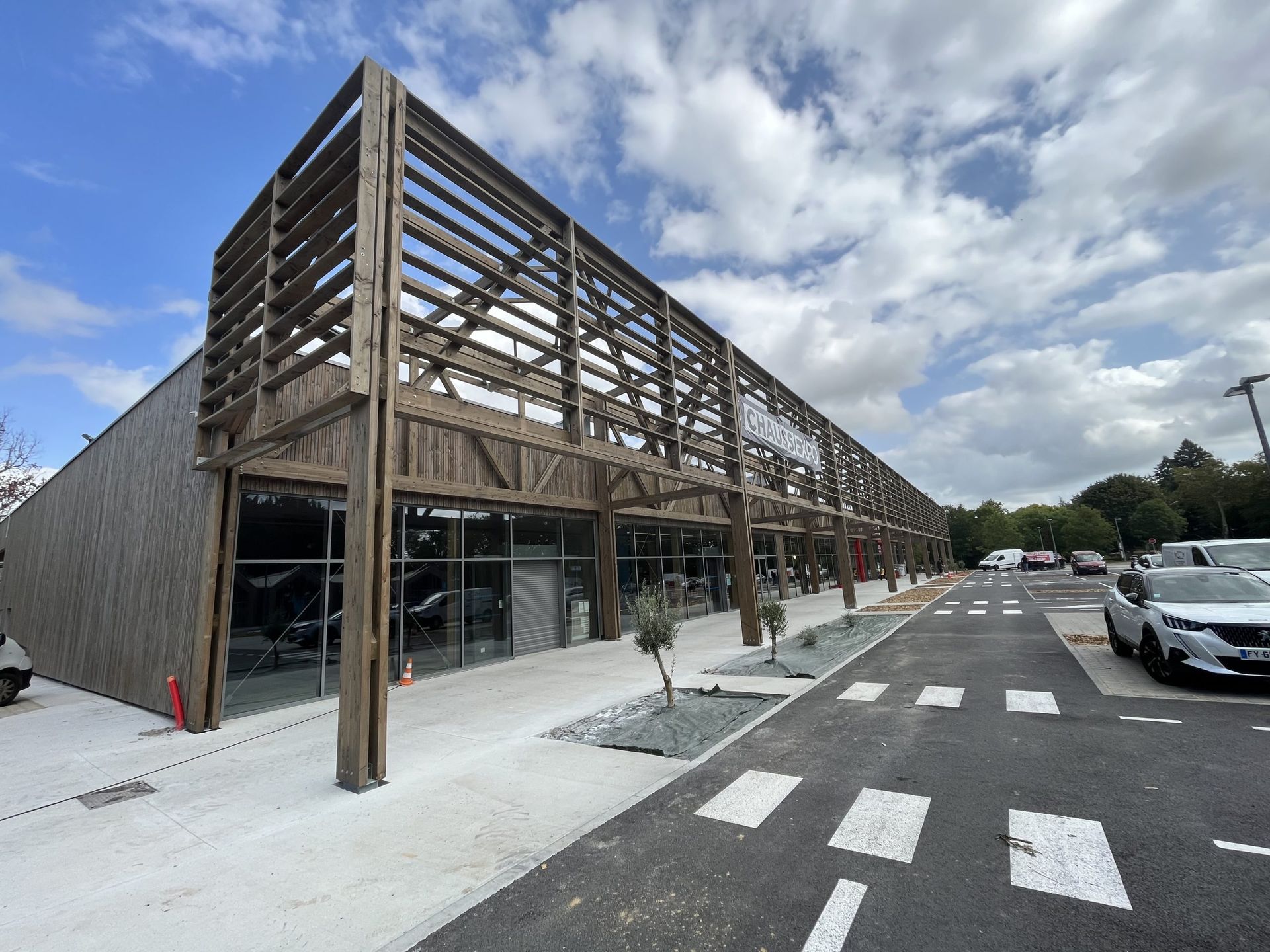 Bâtiment commercial à ossature bois avec de grandes fenêtres et un étage supérieur à ciel ouvert, parking en asphalte gris et ciel nuageux.