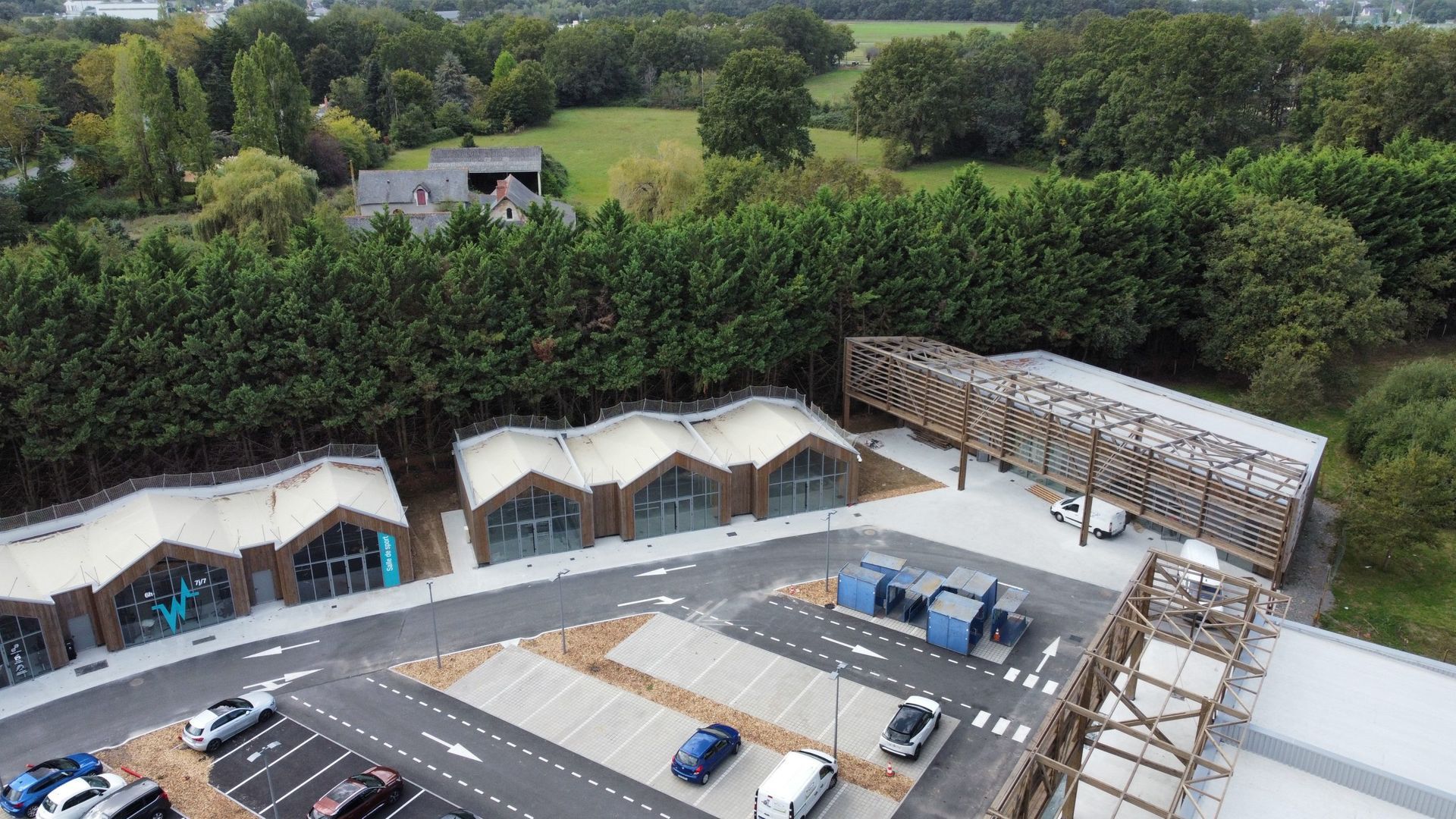 Vue aérienne de bâtiments modernes aux toits voûtés, d'un pont en bois et d'un parking entouré d'arbres.