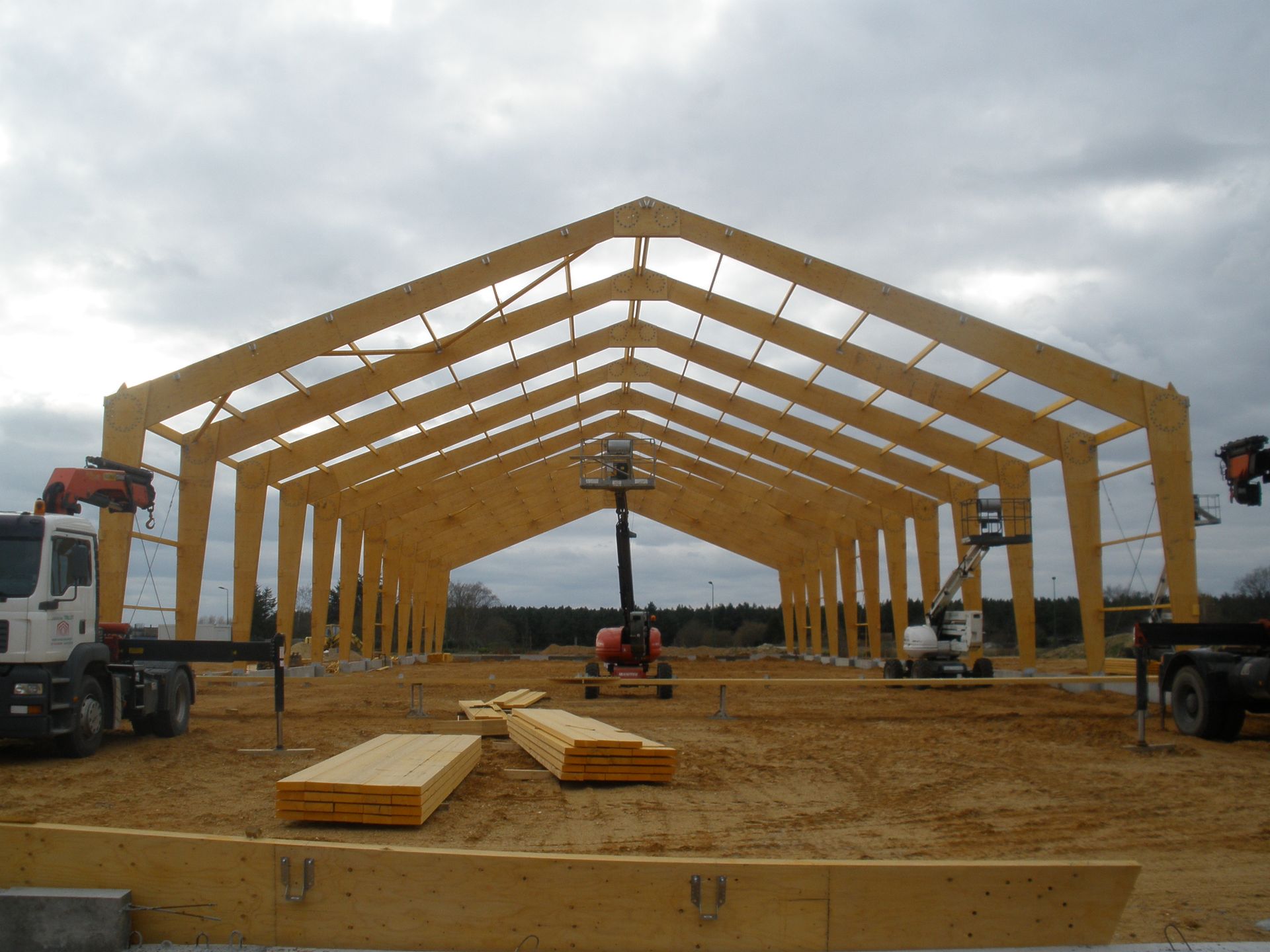 Charpente en bois en construction avec grues, poutres et excavatrice sur un terrain en terre.