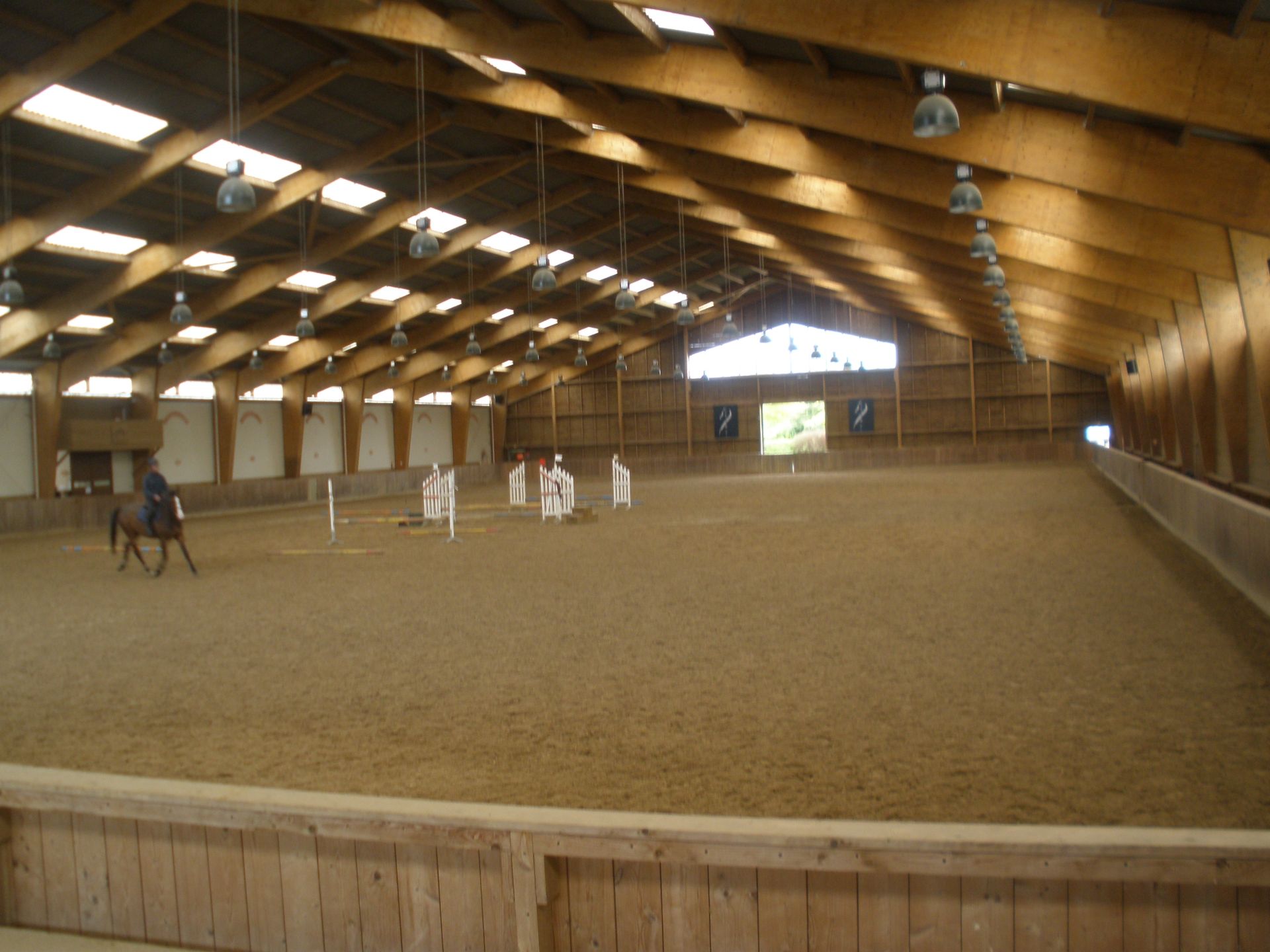 Manège équestre couvert avec un cheval et son cavalier, plusieurs obstacles et un haut plafond en bois.