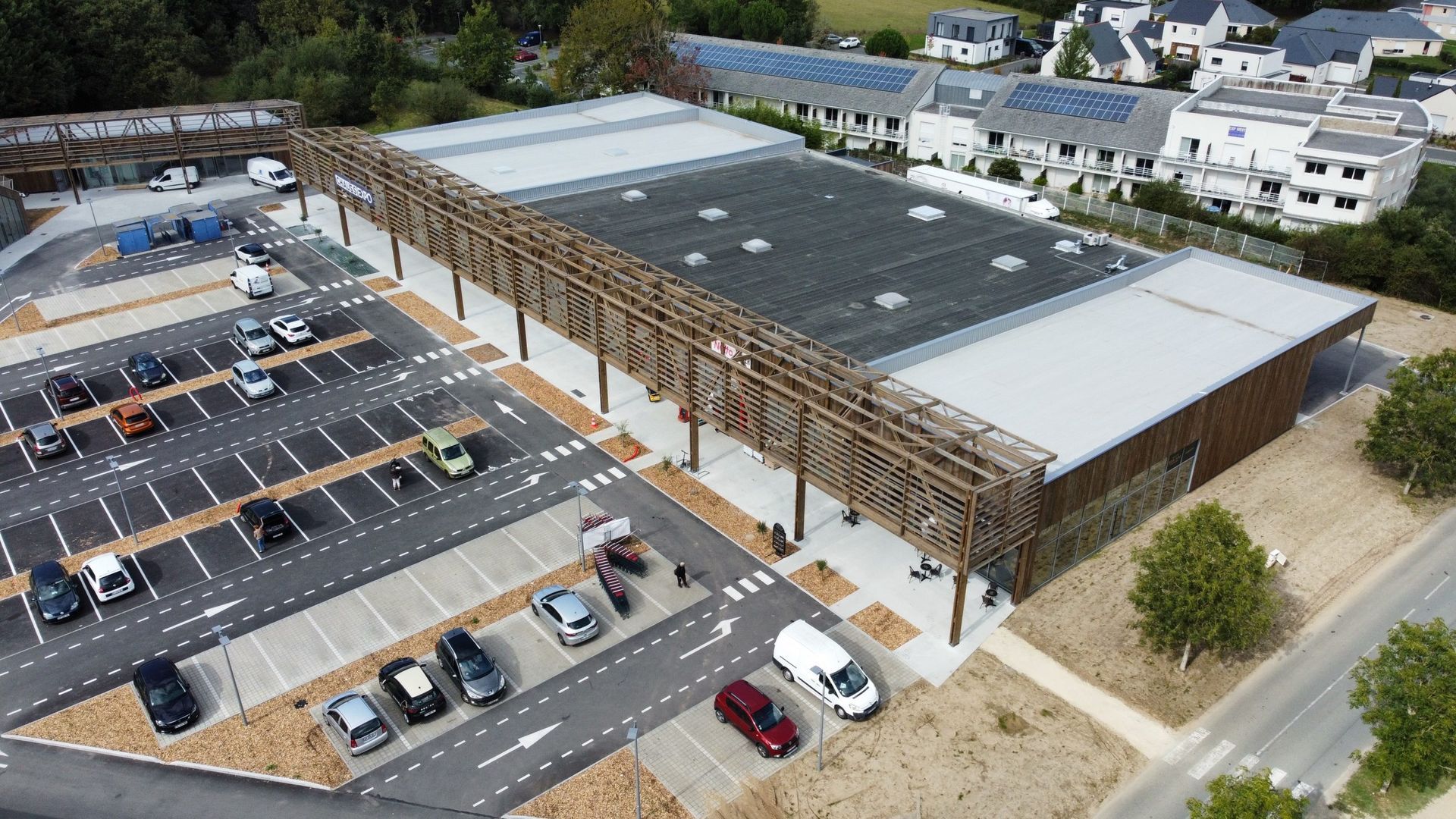 Vue aérienne d'un bâtiment moderne avec une façade en bois, un grand parking et une zone résidentielle en arrière-plan.