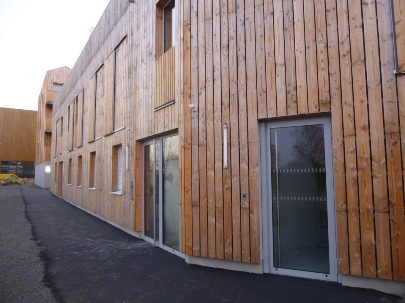 Bâtiment revêtu de bois avec de nombreuses fenêtres et une porte vitrée ; allée piétonne en asphalte devant.