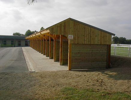 Des abris en bois pour chevaux étaient alignés le long d'un chemin pavé, avec un bâtiment en arrière-plan.