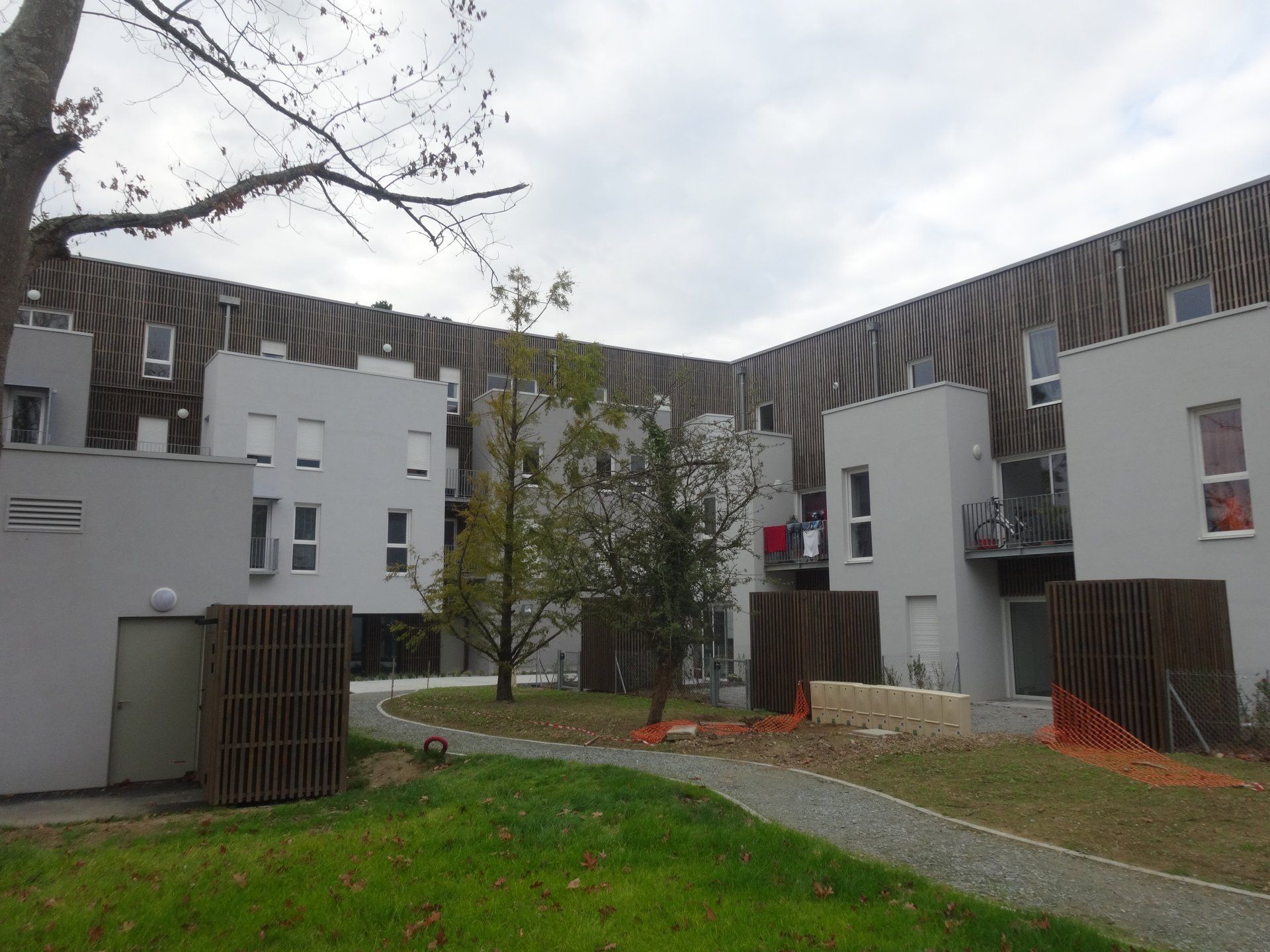 Immeubles d'appartements gris avec des accents de lattes de bois entourant une cour. Ciel couvert.
