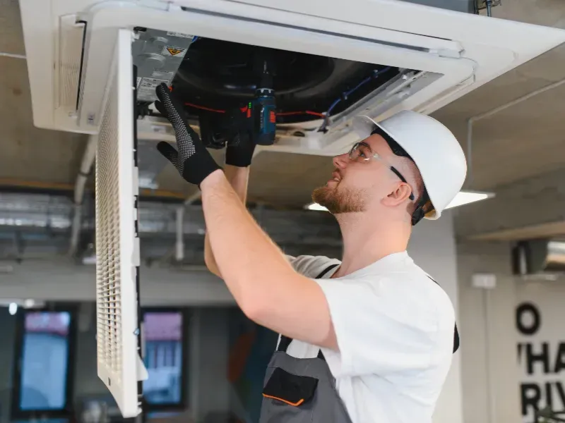 Técnico de HVAC con casco blanco y guantes trabajando en una unidad de aire acondicionado montada en el techo.
